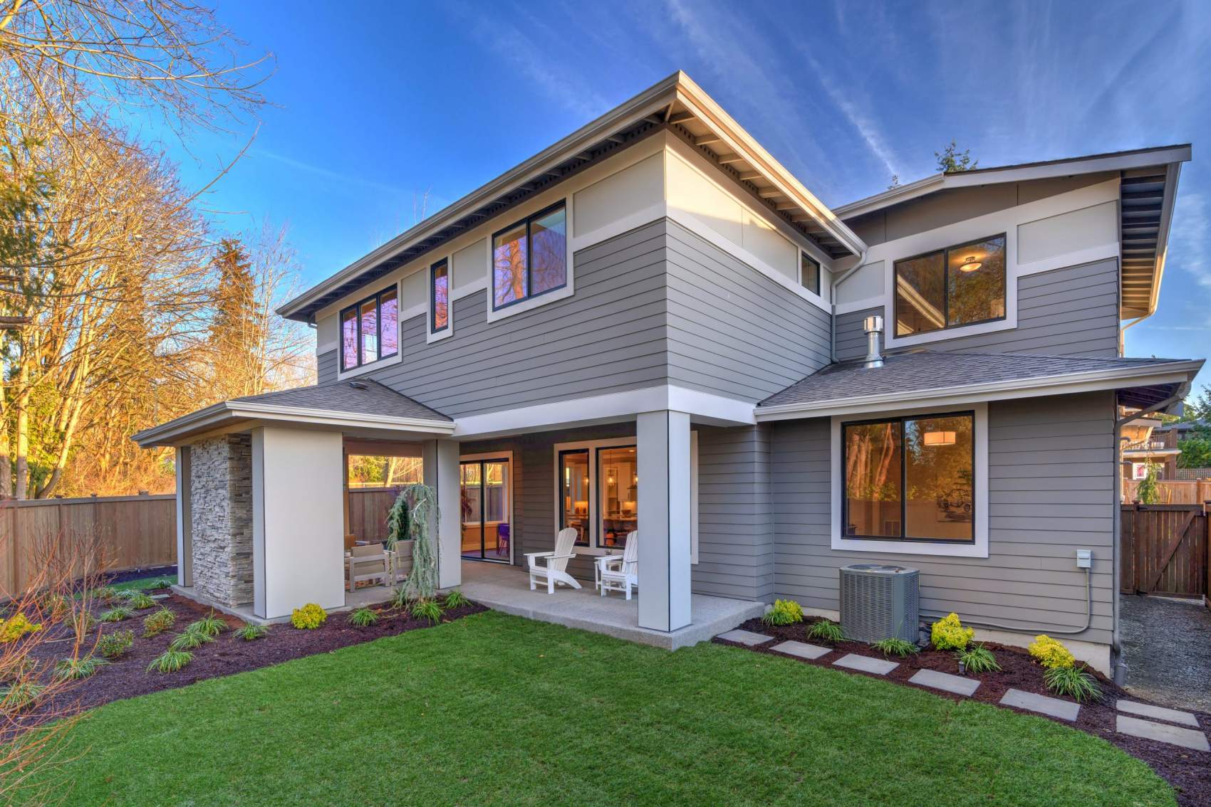 Large modern grey new house with back porch from back yard with green grass and nice landscaping. AC unit, stone elements.