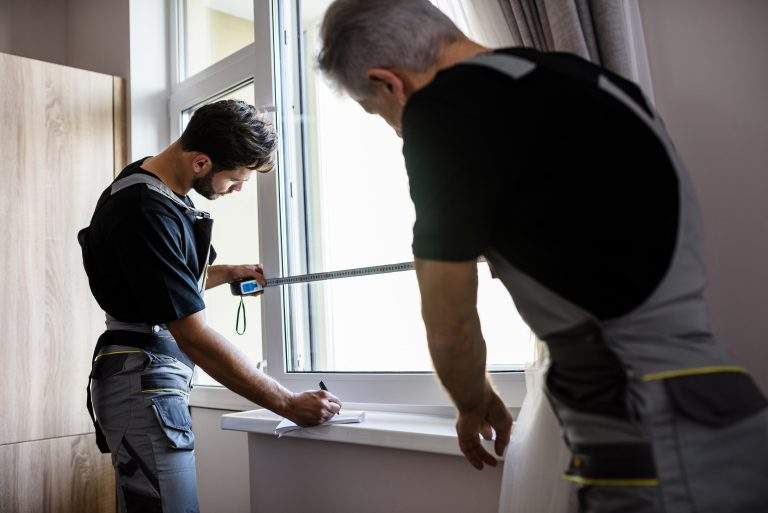 Two professional workers in uniform using tape measure while measuring window and making notes for installing blinds indoors. Construction and maintenance concept. Focus on young man. Horizontal shot